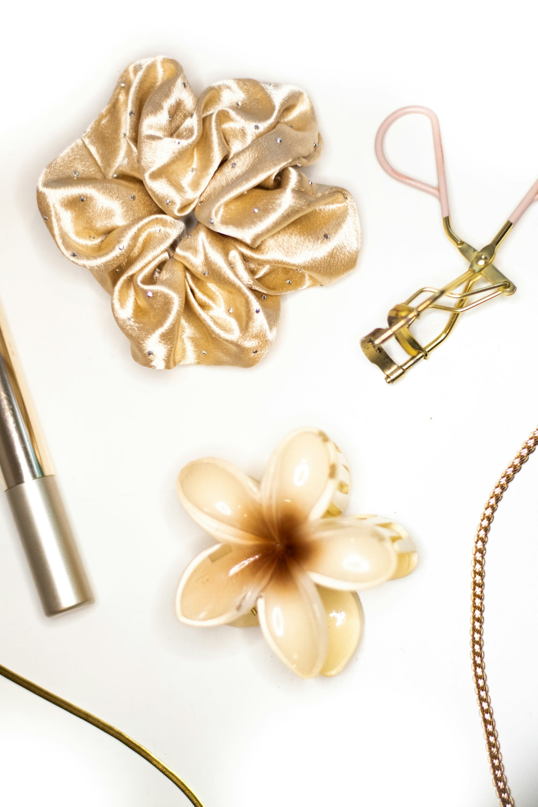 A gold scrunchie, eyelash curler, and flower hair clip on a white surface