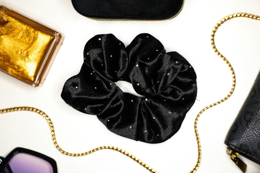 A black velvet scrunchie with small rhinestones beside gold accessories on a white surface