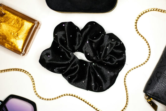 A black velvet scrunchie with small rhinestones beside gold accessories on a white surface
