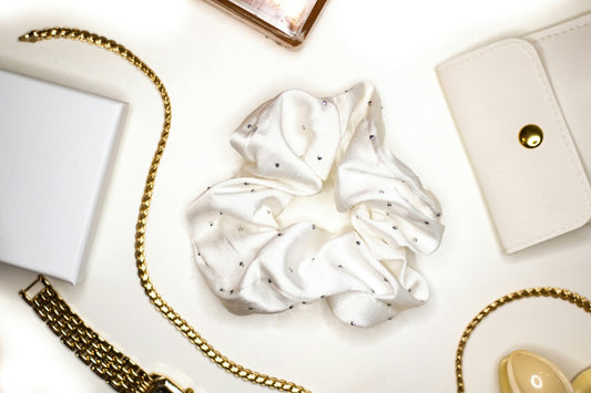A white satin scrunchie with small crystals surrounded by gold jewelry and accessories